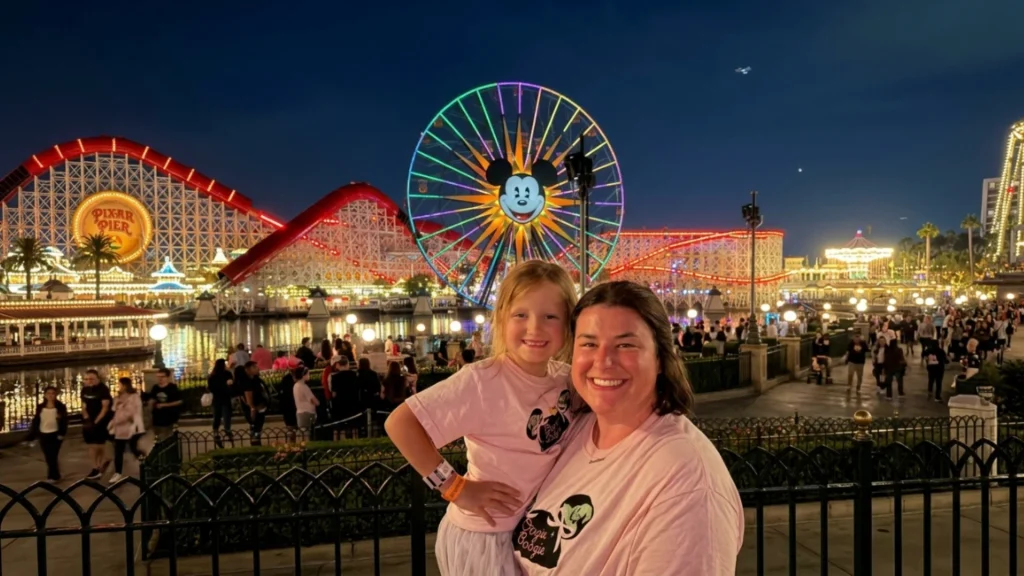 Mother and young daughter smiling at Pixar Pier during nighttime with Pixar Pal-A-Round Ferris wheel lit up at Disney California Adventure